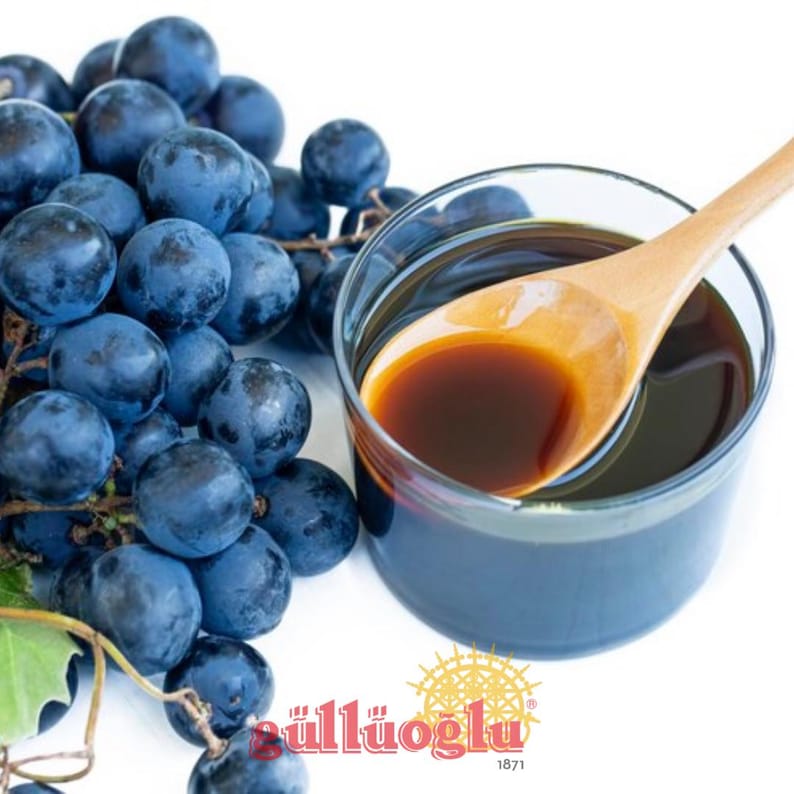 May include: A cluster of dark blue grapes beside a glass of dark liquid with a wooden spoon. The grapes are on a vine, and the liquid appears to be a syrup or juice. The image has a bright, clean background.