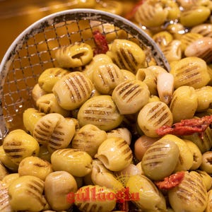 Può includere: Primo piano di olive verdi grigliate con segni di griglia marroni, alcune con noccioli visibili. Le olive sono in una paletta di rete metallica, con alcuni pomodori secchi rossi. Le olive sono di colore verde-giallo chiaro.