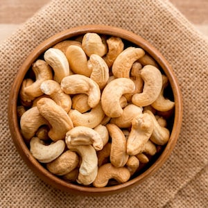 以下が含まれることがあります： 生の Cashew ナッツが入った木製のボウル。 Cashew ナッツは薄茶色で、湾曲した形をしています。 ボウルはジュートの布の上に置かれています。