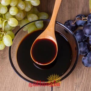 May include: A wooden spoon in a glass bowl containing a dark, thick liquid, possibly a reduction or syrup. Bunches of green and purple grapes are arranged around the bowl, indicating a grape-based product. The background is wooden.