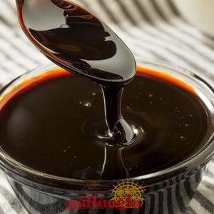 May include: A close-up of dark molasses being poured from a spoon into a clear glass bowl. The molasses is a deep brown with a reddish-brown rim. The bowl sits on a black and white striped cloth.