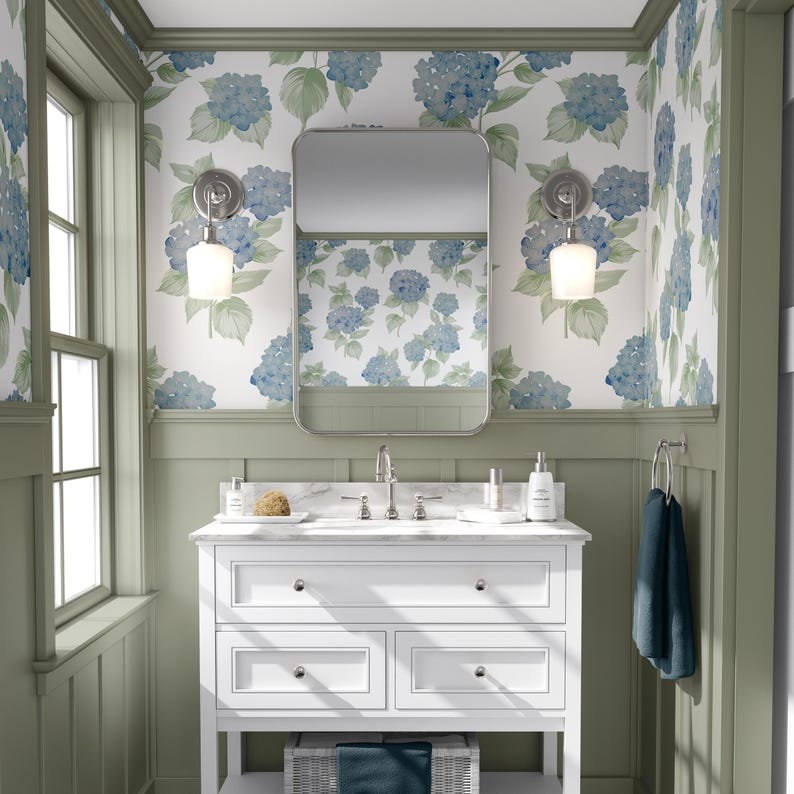 Pu&ograve; includere: Un mobile da bagno bianco con un piano in marmo e tre cassetti. Il mobile &egrave; di fronte a una parete con boiserie verde e carta da parati con ortensie blu. Due applique a parete con paralumi in vetro bianco sono su entrambi i lati di uno specchio sopra il mobile.