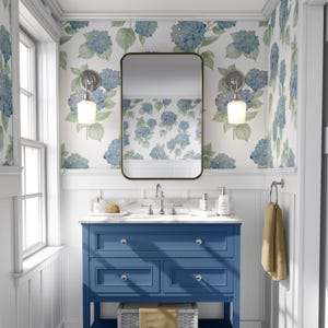 May include: A blue bathroom vanity with three drawers and a white countertop. The vanity has a mirror above it and two wall sconces with white glass shades. The walls are covered in a white wallpaper with a blue floral pattern.