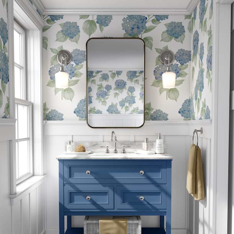 Pu&ograve; includere: Un mobile da bagno blu con tre cassetti e un piano in marmo bianco. Il mobile ha uno specchio sopra e due applique a parete con paralumi in vetro bianco. Le pareti sono rivestite con una carta da parati bianca con un motivo floreale blu.