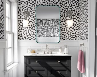 May include: A black and white polka dot wallpaper with a black vanity with a white countertop and a green framed mirror. The vanity has two drawers and a lower shelf with a wicker basket. A pink towel hangs on a towel rack.