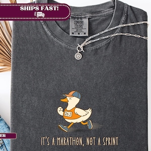 Puede incluir: Camiseta gris oscuro con un pato de dibujos animados que lleva un dorsal con el número 26.2 y una gorra de béisbol. El texto "It's a marathon, not a sprint" está impreso debajo. Un collar plateado está sobre la camiseta.