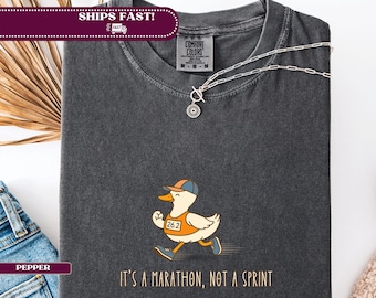 May include: Dark gray t-shirt featuring a cartoon duck wearing a running bib with the number 26.2 and a baseball cap. The text "It's a marathon, not a sprint" is printed below the duck. A silver necklace is draped on the shirt.