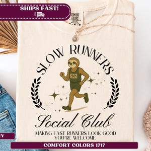 May include: Ivory t-shirt featuring a running sloth graphic wearing a jersey numbered 2605. The shirt displays the text "SLOW RUNNERS Social Club" and "MAKING FAST RUNNERS LOOK GOOD YOU'RE WELCOME".