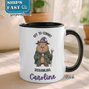 May include: White ceramic mug with a black handle and interior rim. The mug features a cartoon capybara dressed as a wizard, with the text "OFF TO COMMIT SHENANIGANS" and a personalized name. The word "Black" is printed on the mug.
