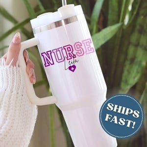 Puede incluir: Vaso t&eacute;rmico blanco con asa, con el texto "NURSE Leila" en morado y rosa, con un s&iacute;mbolo de coraz&oacute;n. El vaso tiene un borde plateado y una tapa blanca. La imagen tambi&eacute;n incluye una pegatina "SHIPS FAST!"