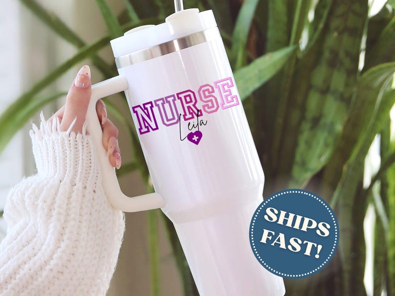 Puede incluir: Vaso t&eacute;rmico blanco con asa, con el texto "NURSE Leila" en morado y rosa, con un s&iacute;mbolo de coraz&oacute;n. El vaso tiene un borde plateado y una tapa blanca. La imagen tambi&eacute;n incluye una pegatina "SHIPS FAST!"