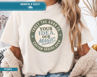 以下が含まれることがあります： 「Trust the Good Co. Your Idea. Our Magic. Custom Orders Open」と書かれた円形グラフィックが特徴のアイボリー色のTシャツ。シャツには「Ships Fast！」のバナーも付いています。