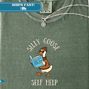 Könnte beinhalten: Moosgrünes T-Shirt mit einer Grafik einer Gans mit Sonnenbrille und Pullover, die ein Buch mit dem Titel "Silly Goose Guide to Being Taken Seriously" liest. Die Worte "Silly Goose" und "Self Help" sind ebenfalls auf dem Shirt aufgedruckt.