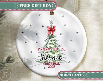 May include: White ceramic Christmas ornament with a hand-drawn Christmas tree design. The tree is adorned with a red bow and the words "PROMOTED TO Nana CHRISTMAS 2025". Black snowflakes are scattered around the tree.