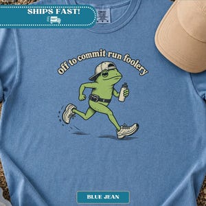 Könnte beinhalten: Ein blaues T-Shirt mit einer Grafik eines grünen Frosches, der eine Baseballkappe und eine Gürteltasche trägt und rennt. Der Frosch hält eine Wasserflasche und der Text darüber lautet off to commit run foolery in geschwungener Schrift.