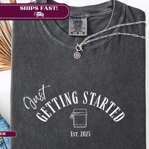以下が含まれることがあります： 「Just Getting Started」の文字と瓶のグラフィックが入った濃いグレーのTシャツ。Tシャツはクルーネックで半袖です。ペンダント付きのシルバーのネックレスがTシャツの上にあります。