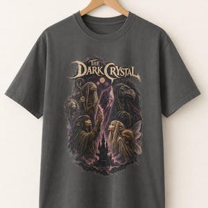 May include: A charcoal grey short-sleeve t-shirt featuring a graphic print of The Dark Crystal. The design displays characters, a glowing crystal, and a castle in shades of purple, brown, and cream, centred on the front of the garment.