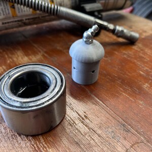 May include: A close-up of a silver wheel bearing and a silver metal cap with a small hole on top. The objects are on a wooden surface.