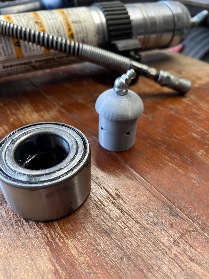 May include: A close-up of a silver wheel bearing and a silver metal cap with a small hole on top. The objects are on a wooden surface.