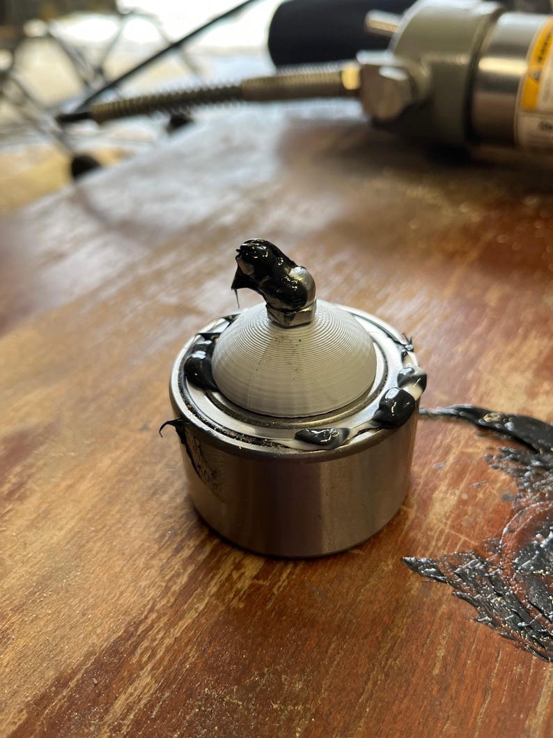 May include: A close-up of a silver bearing with a black rubber seal and a black grease coating. The bearing is sitting on a wooden surface.