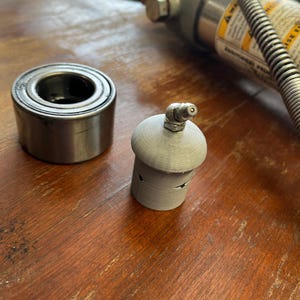 May include: A close-up of a silver bearing race and a gray 3D printed bearing race installer tool. The tool has a small hole in the top and a small hole on the side.