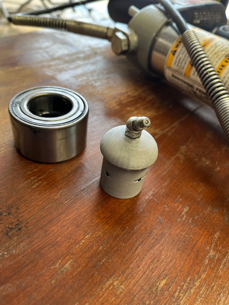 May include: A close-up of a silver bearing race and a gray 3D printed bearing race installer tool. The tool has a small hole in the top and a small hole on the side.