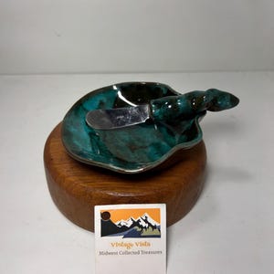 May include: A teal and brown ceramic butter dish with a stainless steel knife, resting on a wooden base. The dish has a unique shape and a glossy finish. The knife is small and has a simple design. The dish is labeled "Vintage Vista Midwest Collected Treasures."
