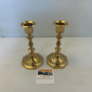 May include: Two vintage brass candlesticks with a hexagonal base and ornate stem. The candlesticks have a polished gold finish and are displayed on a white surface. A small card with the text "Vintage Vista Midwest Collected Treasures" is in the foreground.
