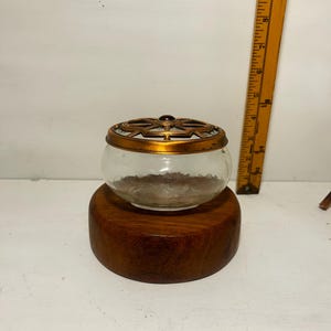May include: A clear glass bowl with a gold-toned metal lid featuring a geometric cutout pattern and a small dark center accent, resting on a thick circular wooden base. A yellow ruler shows the object height is approximately 3.5 inches.