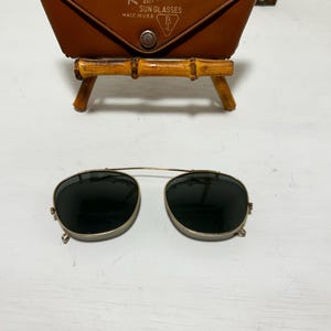 Op de afbeelding: Vintage Ray-Ban zonnebril met een bruine leren etui. De zonnebril heeft een goudkleurig montuur en donkere glazen. De etui is voorzien van het Ray-Ban logo en de tekst "SUN GLASSES MADE IN U.S.A."