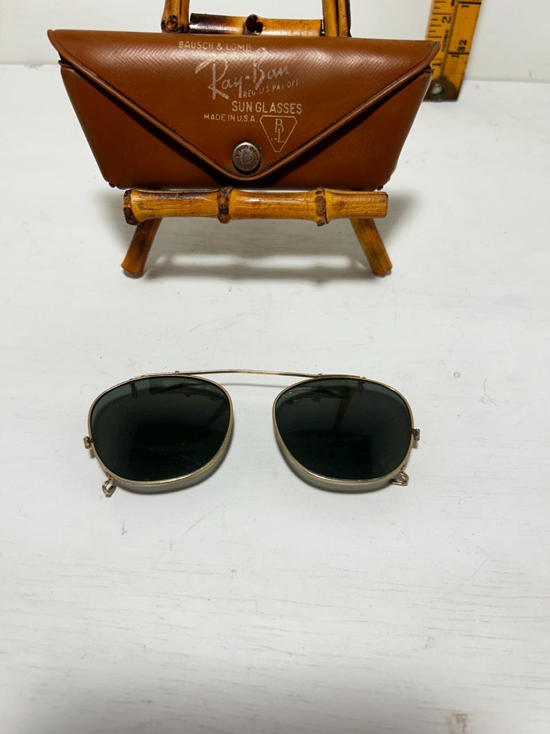 Op de afbeelding: Vintage Ray-Ban zonnebril met een bruine leren etui. De zonnebril heeft een goudkleurig montuur en donkere glazen. De etui is voorzien van het Ray-Ban logo en de tekst "SUN GLASSES MADE IN U.S.A."