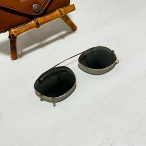 Op de afbeelding: Vintage Ray-Ban zonnebril met donkere glazen en een goudkleurig montuur. De zonnebril ligt naast een bruine leren etui met het Ray-Ban logo en tekst. De etui heeft een bamboe standaard.