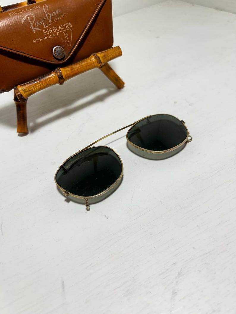 Op de afbeelding: Vintage Ray-Ban zonnebril met donkere glazen en een goudkleurig montuur. De zonnebril ligt naast een bruine leren etui met het Ray-Ban logo en tekst. De etui heeft een bamboe standaard.