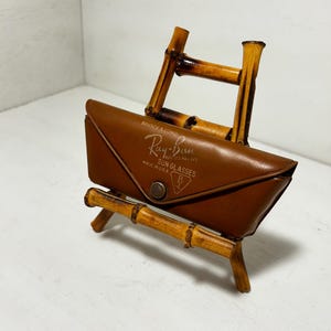 Op de afbeelding: Een bruin leren Ray-Ban zonnebril etui met merknaam en logo, op een bamboe standaard. De etui is rechthoekig met een druksluiting. De standaard is gemaakt van bamboestokken.