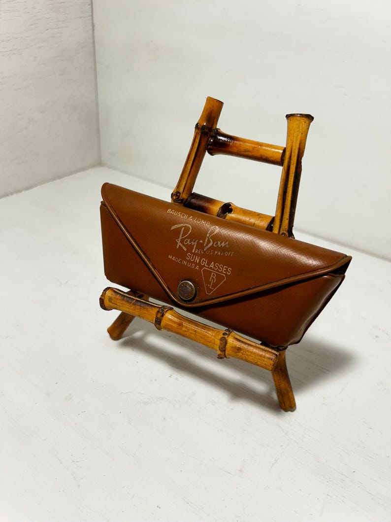Op de afbeelding: Een bruin leren Ray-Ban zonnebril etui met merknaam en logo, op een bamboe standaard. De etui is rechthoekig met een druksluiting. De standaard is gemaakt van bamboestokken.
