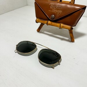 Op de afbeelding: Vintage Ray-Ban zonnebril met een bruine leren etui. De zonnebril heeft een goudkleurig montuur en donkere glazen. De etui is voorzien van het Ray-Ban logo en de tekst "SUN GLASSES MADE IN U.S.A."