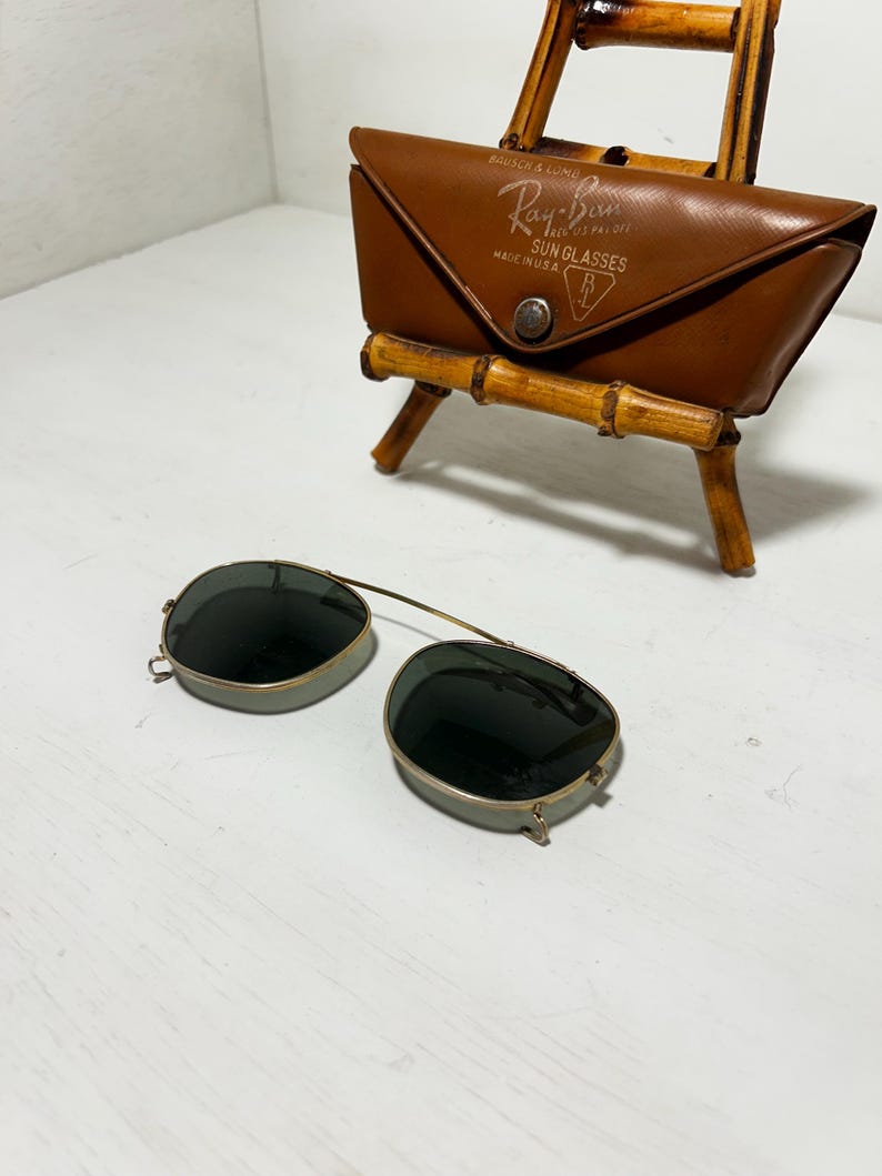 Op de afbeelding: Vintage Ray-Ban zonnebril met een bruine leren etui. De zonnebril heeft een goudkleurig montuur en donkere glazen. De etui is voorzien van het Ray-Ban logo en de tekst "SUN GLASSES MADE IN U.S.A."