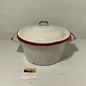 May include: Vintage white enamelware pot with a lid. The pot has a red trim and handles. The lid has a small handle. The pot is likely used for cooking. The pot is a vintage item.