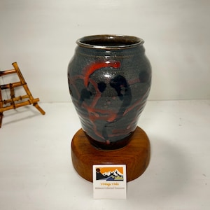 May include: A dark gray ceramic vase with red and black abstract designs. The vase sits on a round wooden base. The rim of the vase has a dark brown color. The vase is a vintage item.