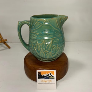 Könnte beinhalten: Eine türkisfarbene Keramikkrug mit Henkel und Ausguss, verziert mit einem erhabenen floralen Design. Der Krug steht auf einem runden Holzsockel. Das Etikett lautet "Vintage Vista Midwest Collected Treasures."