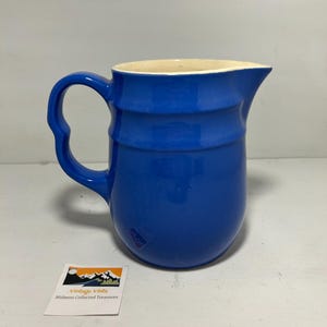 Op de afbeelding: Een kobaltblauwe keramische kan met een afgeronde body en een gebogen handvat. De kan heeft een crèmekleurig interieur en een tuit om uit te schenken. De woorden "Vintage Vista Midwest Collected Treasures" staan op een kleine kaart.