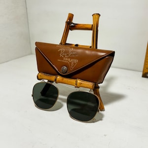 Op de afbeelding: Vintage Ray-Ban zonnebril met een bruine leren etui, tentoongesteld op een bamboe standaard. De etui heeft het Ray-Ban logo en tekst. De zonnebril heeft vierkante, donkergroene glazen en een zilveren montuur.