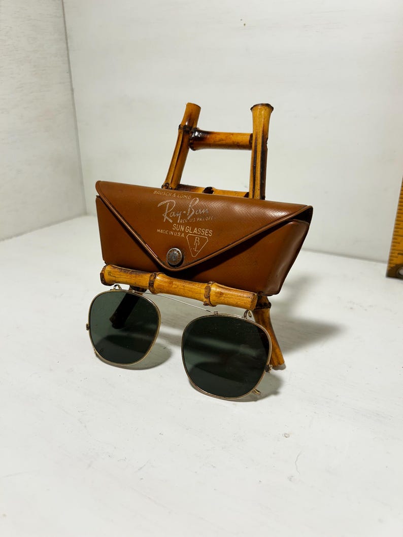 Op de afbeelding: Vintage Ray-Ban zonnebril met een bruine leren etui, tentoongesteld op een bamboe standaard. De etui heeft het Ray-Ban logo en tekst. De zonnebril heeft vierkante, donkergroene glazen en een zilveren montuur.