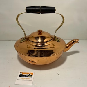 May include: A copper tea kettle with a black wooden handle and brass accents. The kettle has a rounded body, a lid with a small knob, and a spout. The handle arches over the top. A small sticker is visible.