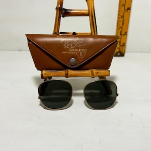 Op de afbeelding: Vintage Ray-Ban zonnebril met een bruine leren etui, tentoongesteld op een bamboe standaard. De etui is voorzien van het Ray-Ban logo en de tekst "MADE IN U.S.A.". De zonnebril heeft donkere glazen en een goudkleurig montuur.