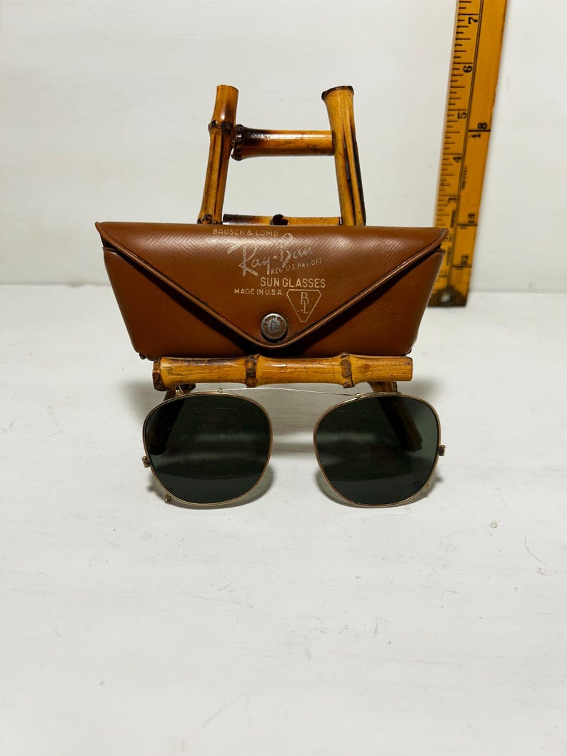 Op de afbeelding: Vintage Ray-Ban zonnebril met een bruine leren etui, tentoongesteld op een bamboe standaard. De etui is voorzien van het Ray-Ban logo en de tekst "MADE IN U.S.A.". De zonnebril heeft donkere glazen en een goudkleurig montuur.