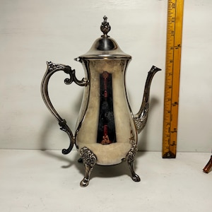 May include: Silver-plated teapot with ornate handle and spout. The teapot has a decorative finial on the lid and rests on three small feet. A ruler is next to the teapot. The teapot has a decorative design on the front.