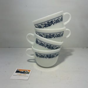 Puede incluir: Una pila de cuatro tazas de té blancas con un diseño floral azul alrededor del borde. Cada taza tiene un asa y están apiladas una encima de la otra. Las tazas están sobre una superficie blanca. Una pequeña tarjeta con texto está en primer plano.