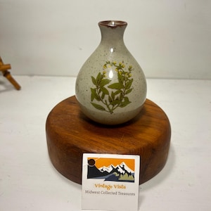 May include: A small, speckled ceramic vase with a brown rim and a botanical design featuring green leaves and yellow flowers. The vase is displayed on a round, wooden base. A small tag with the text "Vintage Vista Midwest Collected Treasures" is in front of the base.