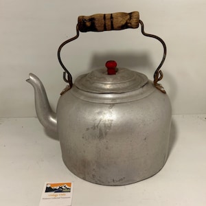 Op de afbeelding: Vintage aluminium waterkoker met een houten handvat en een rode knop op het deksel. De waterkoker heeft een tuit en een metalen handvat. De waterkoker is licht zilverkleurig en vertoont gebruikssporen.